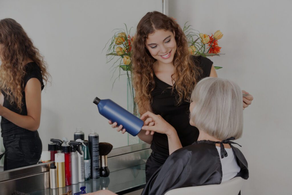 Une jeune coiffeuse souriante aux cheveux bouclés tient un flacon de soin capillaire bleu tout en conseillant une femme âgée aux cheveux argentés assise dans un fauteuil de salon.