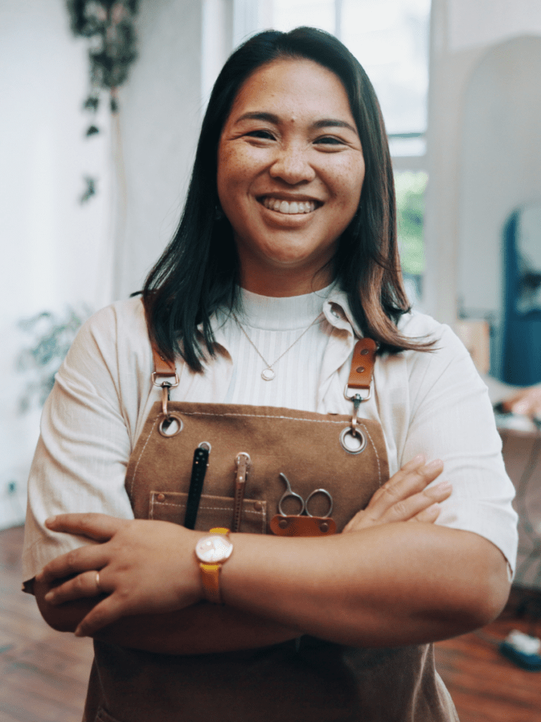 Portrait d'une professionnelle de salon souriante aux cheveux foncés, regardant l'objectif les bras croisés. Elle porte une chemise claire sous un tablier en toile marron équipé de pinces à cheveux et de ciseaux.