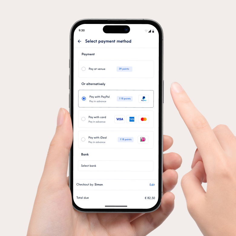 Gros plan d'un smartphone affichant l'interface de paiement de l'application Treatwell pour sélectionner un mode de paiement. Les options incluent le paiement sur place ou le paiement anticipé avec PayPal, carte ou iDeal. Une main tient le téléphone et un doigt pointe vers l'écran.