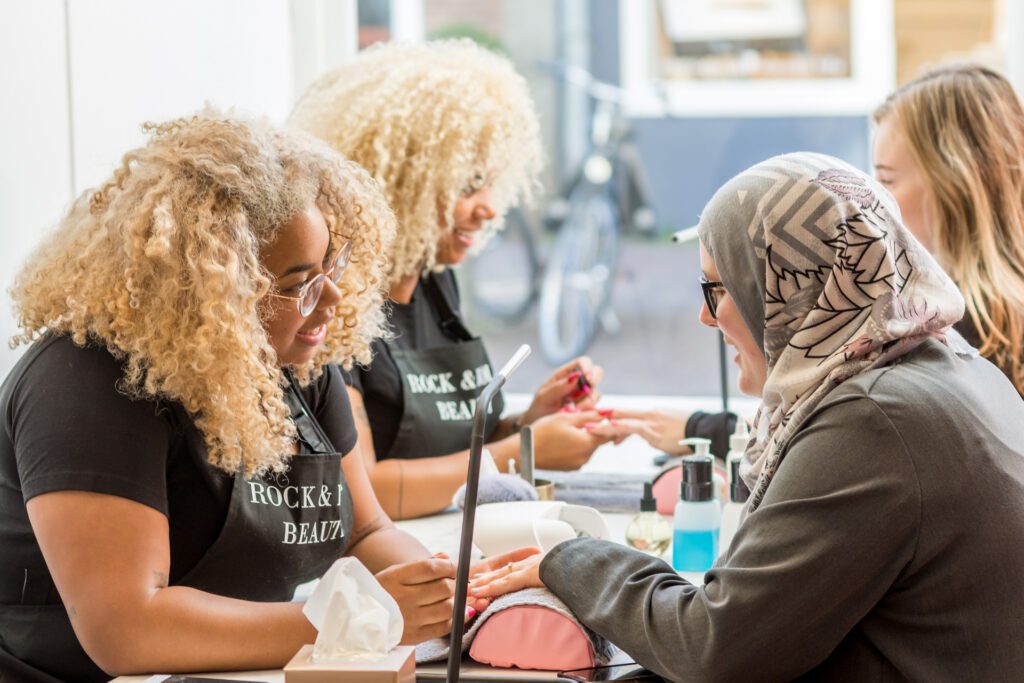 Deux manicures aux cheveux bouclés blonds portant des tabliers noirs avec l'inscription "Rock & Roll Beauty" font une manucure à des clientes sur une longue table. Une cliente porte un hijab gris.