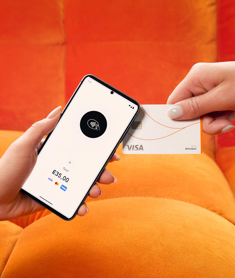 Une main tient un smartphone faisant office de terminal de paiement, affichant une icône de paiement sans contact et un total de 35,00 £, tandis qu'une autre main approche une carte de crédit Visa blanche pour effectuer le paiement. L'arrière-plan est un canapé orange vif.