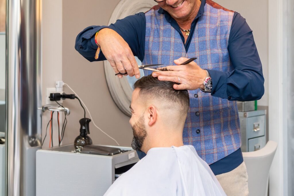 Un coiffeur portant un gilet à carreaux bleu coupe les cheveux d'un client avec des ciseaux et un peigne dans un salon. Le client a les cheveux courts, une barbe et porte un peignoir de coiffure blanc.