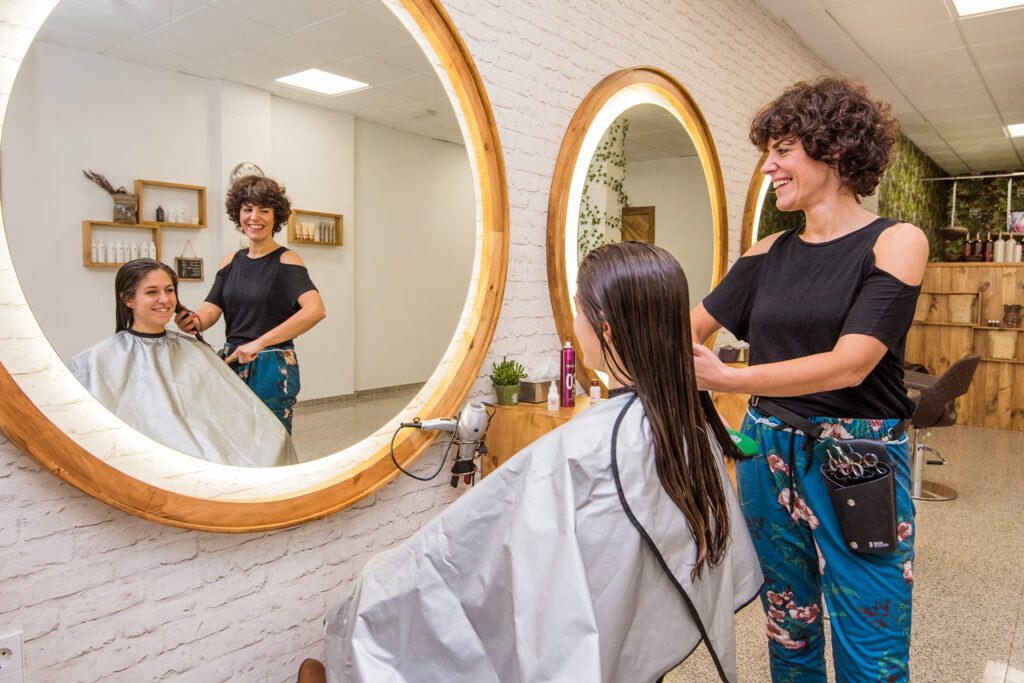 Une coiffeuse souriante aux cheveux courts bouclés brosse les longs cheveux humides d'une cliente devant un grand miroir rond éclairé. La scène se reflète dans le miroir, montrant l'intérieur du salon.