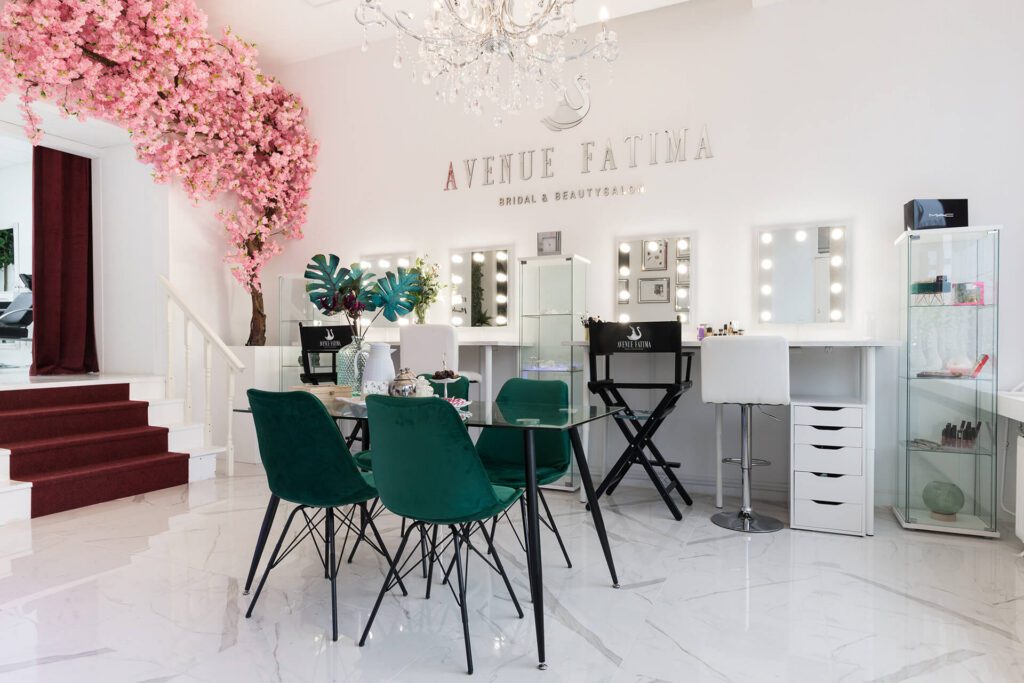 Un salon de beauté et de coiffure de mariée lumineux et élégant nommé "Avenue Fatima". L'espace comprend plusieurs postes de maquillage avec miroirs éclairés, des chaises en velours vert émeraude autour d'une table en verre et un cerisier rose décoratif.