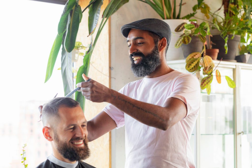 Un coiffeur souriant avec une barbe foncée et une casquette plate grise utilise des ciseaux et un peigne sur les cheveux d'un client souriant dans un salon lumineux décoré de plantes vertes.