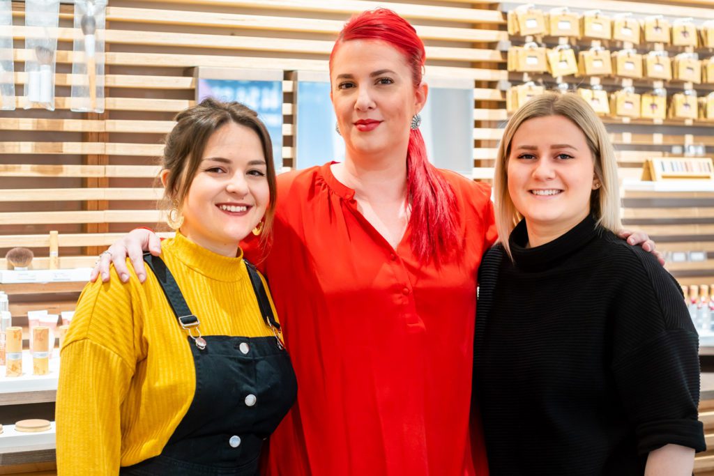Trois professionnelles de salon souriantes posent ensemble, bras dessus bras dessous, devant un mur à lattes en bois présentant des cosmétiques. La femme au centre a des cheveux rouge vif et un haut rouge.