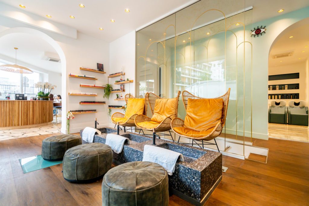 L'intérieur élégant et lumineux d'un salon de beauté doté d'un espace pédicure avec trois fauteuils en osier jaune moutarde, des bacs en pierre avec des serviettes blanches et des repose-pieds gris assortis sur un parquet.