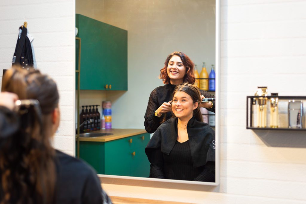 Une coiffeuse souriante aux cheveux auburn coiffe les cheveux d'une cliente souriante avec un lisseur, vue à travers le reflet d'un grand miroir rectangulaire dans un salon moderne aux meubles vert émeraude.