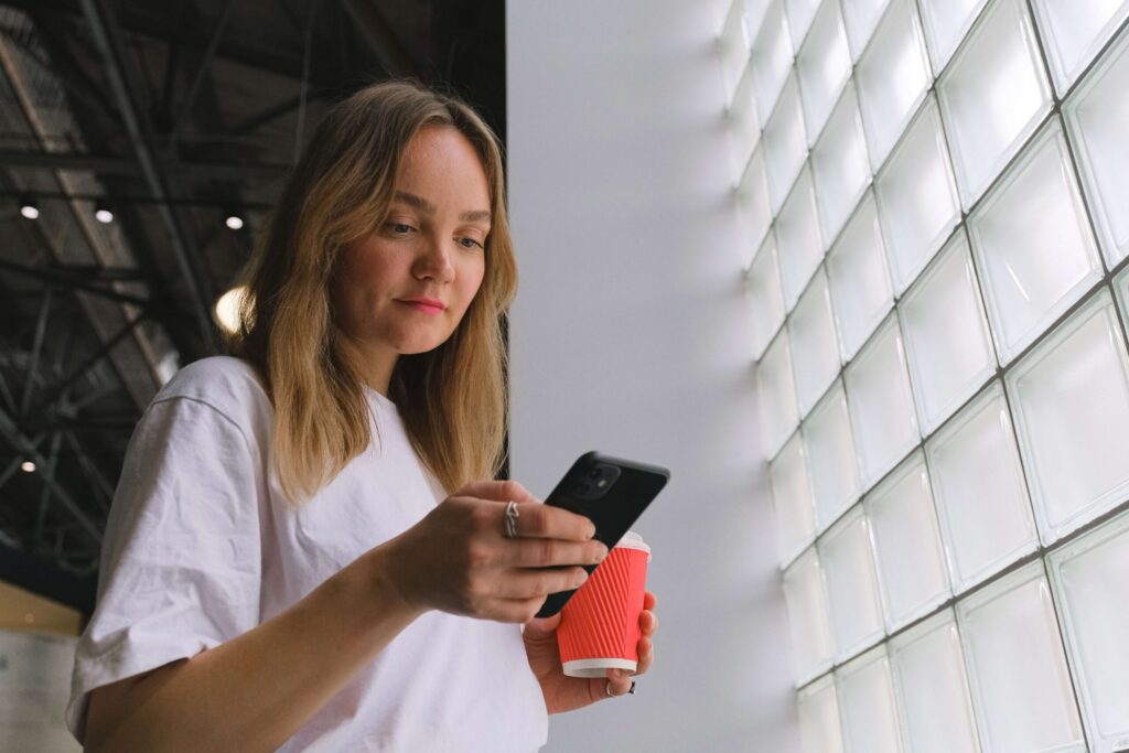 Une vue en contre-plongée d'une jeune femme aux cheveux blonds, portant un t-shirt blanc, qui regarde son smartphone noir tout en tenant un gobelet de café à emporter rouge. Elle se tient à côté d'un grand mur de briques de verre.