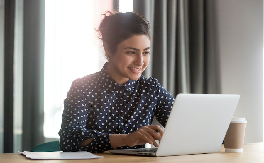 Une femme souriante avec un chignon, portant une chemise bleu foncé à pois, est assise à un bureau en bois et tape sur un ordinateur portable argenté à côté d'un gobelet de café à emporter.