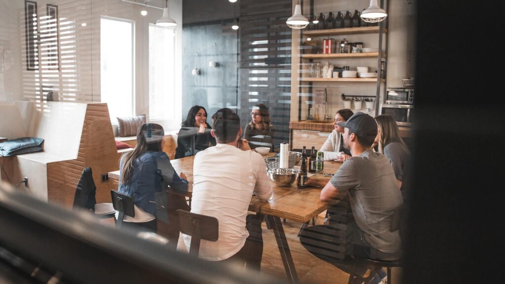 Une vue à travers une vitre d'un groupe de collègues assis autour d'une longue table en bois dans une cuisine ou salle à manger de bureau moderne, discutant et partageant des boissons.