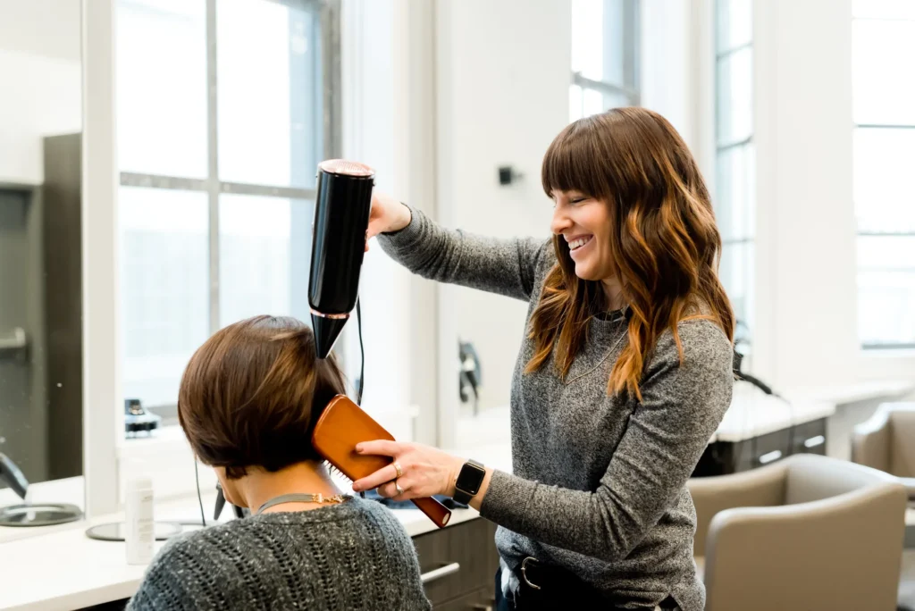 Une coiffeuse souriante réalise un brushing sur les cheveux d'un client dans un salon lumineux et moderne.